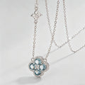 A silver-toned double-layer necklace featuring a main flower pendant with light blue gemstones and a smaller clover charm on a delicate chain, set against a clean light gray background.