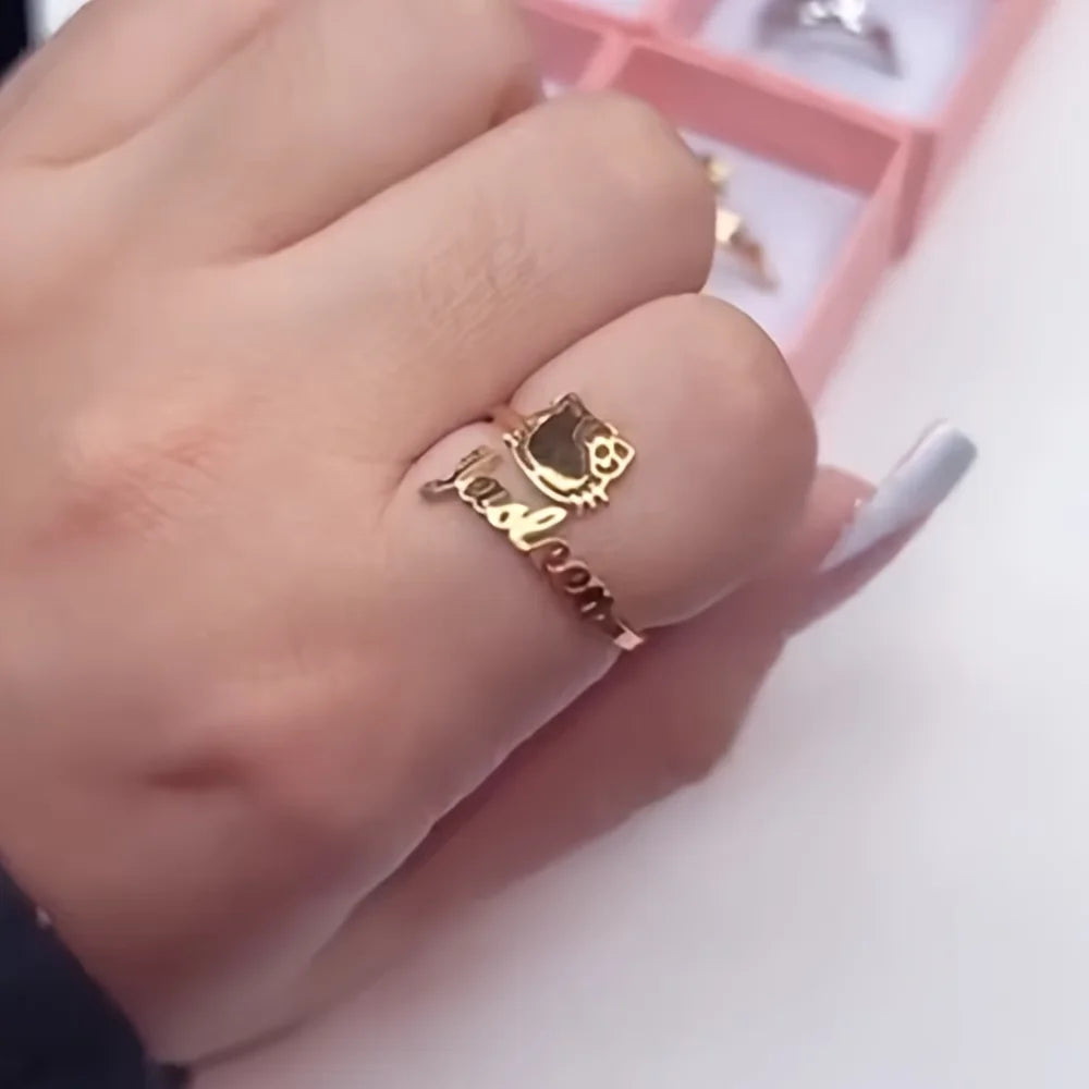 A close-up of a hand wearing a gold ring with a Hello Kitty face and the phrase 'just dream' in cursive script. A pink ring box is visible in the blurred background.
