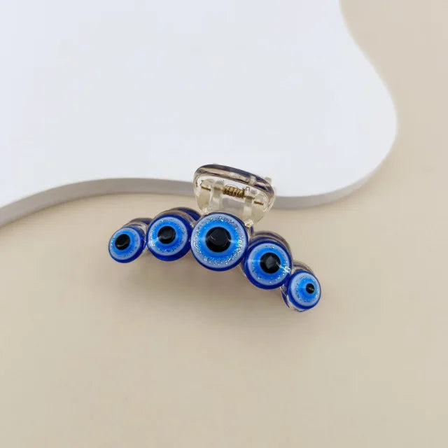 A clear plastic claw hair clip decorated with five blue and white evil eye beads, shown on a neutral beige background.