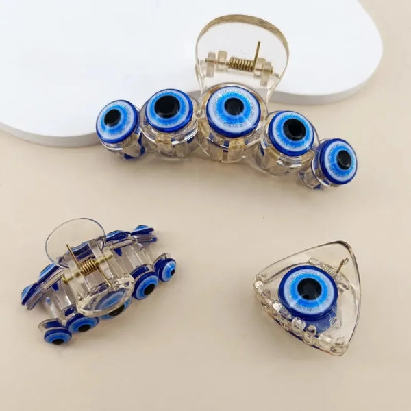 Three clear acrylic hair accessories with blue and black evil eye designs, including a large claw clip, a smaller claw clip, and a triangular barrette, arranged on a light beige surface.