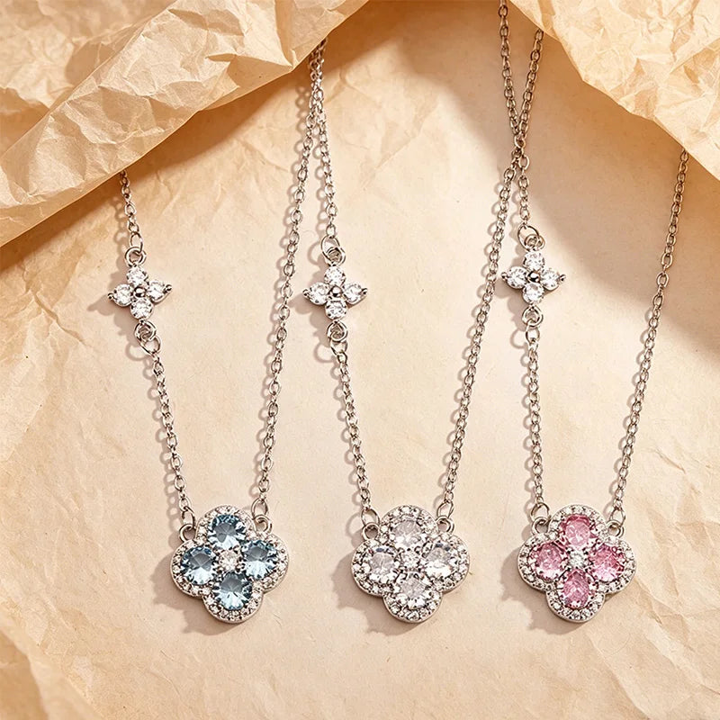 Three silver-tone floral pendant necklaces with clear and colored gemstones are arranged on a crumpled beige paper background. The left necklace has a light blue flower pendant, the middle has a clear flower, and the right has a pink flower.