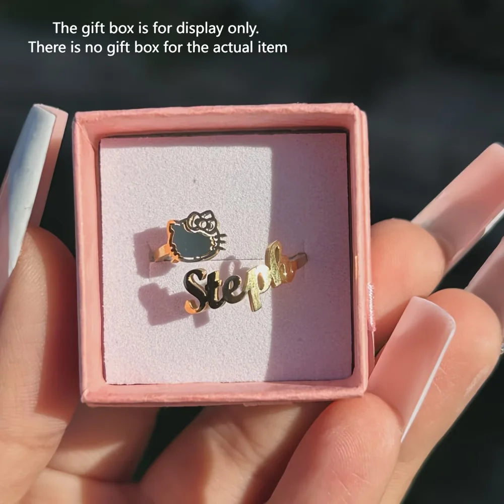 A pair of gold-toned earrings displayed in a pink presentation box held by hands with long manicured nails. One earring features a Hello Kitty face with a dark green enamel patch, and the other is a gold script nameplate reading 'Steph'.