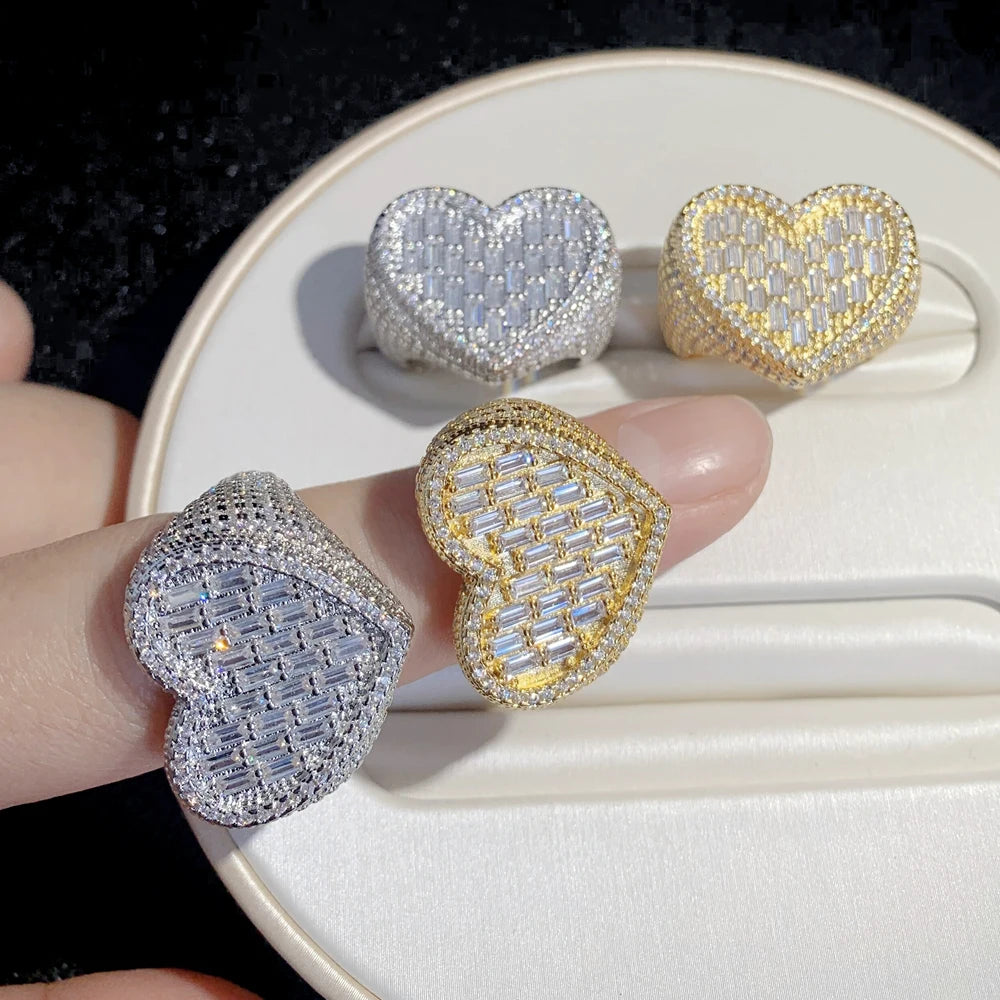 Four heart-shaped pave rings displayed on a white ring box, with one ring being worn on a finger. The rings feature silver and gold settings with clear baguette and round stones.