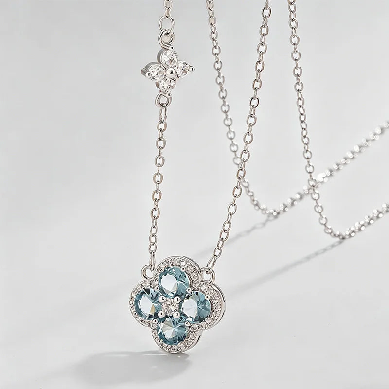A silver-toned double-layer necklace featuring a main flower pendant with light blue gemstones and a smaller clover charm on a delicate chain, set against a clean light gray background.